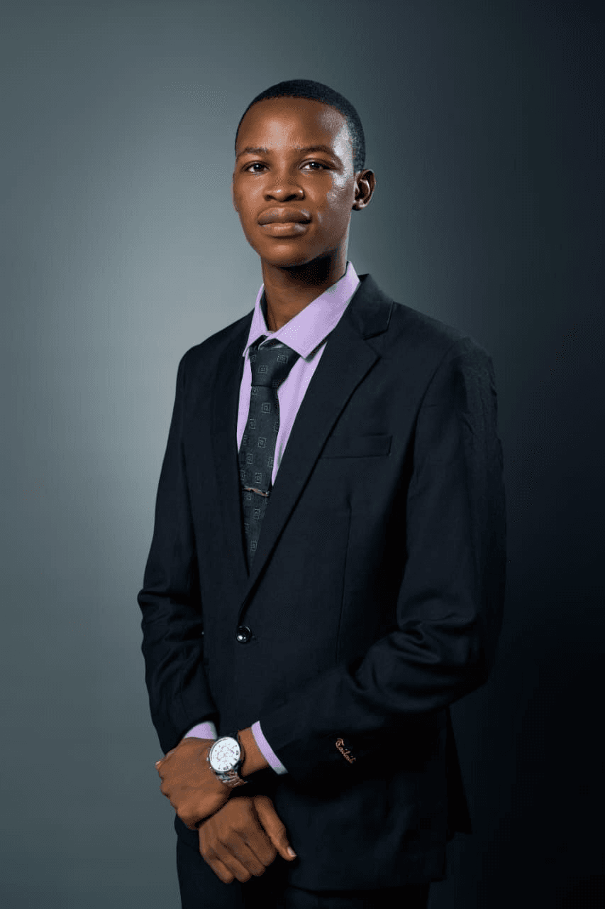 Timothy Adejare Ayelagbe, in a dark suit and lilac shirt, photographed against a grey studio background.